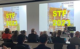 A photo taken from behind a seated audience of people. They are looking towards a woman who is presenting and on the dual screens we can see the words STF 2023 ' Beyond the Code'.